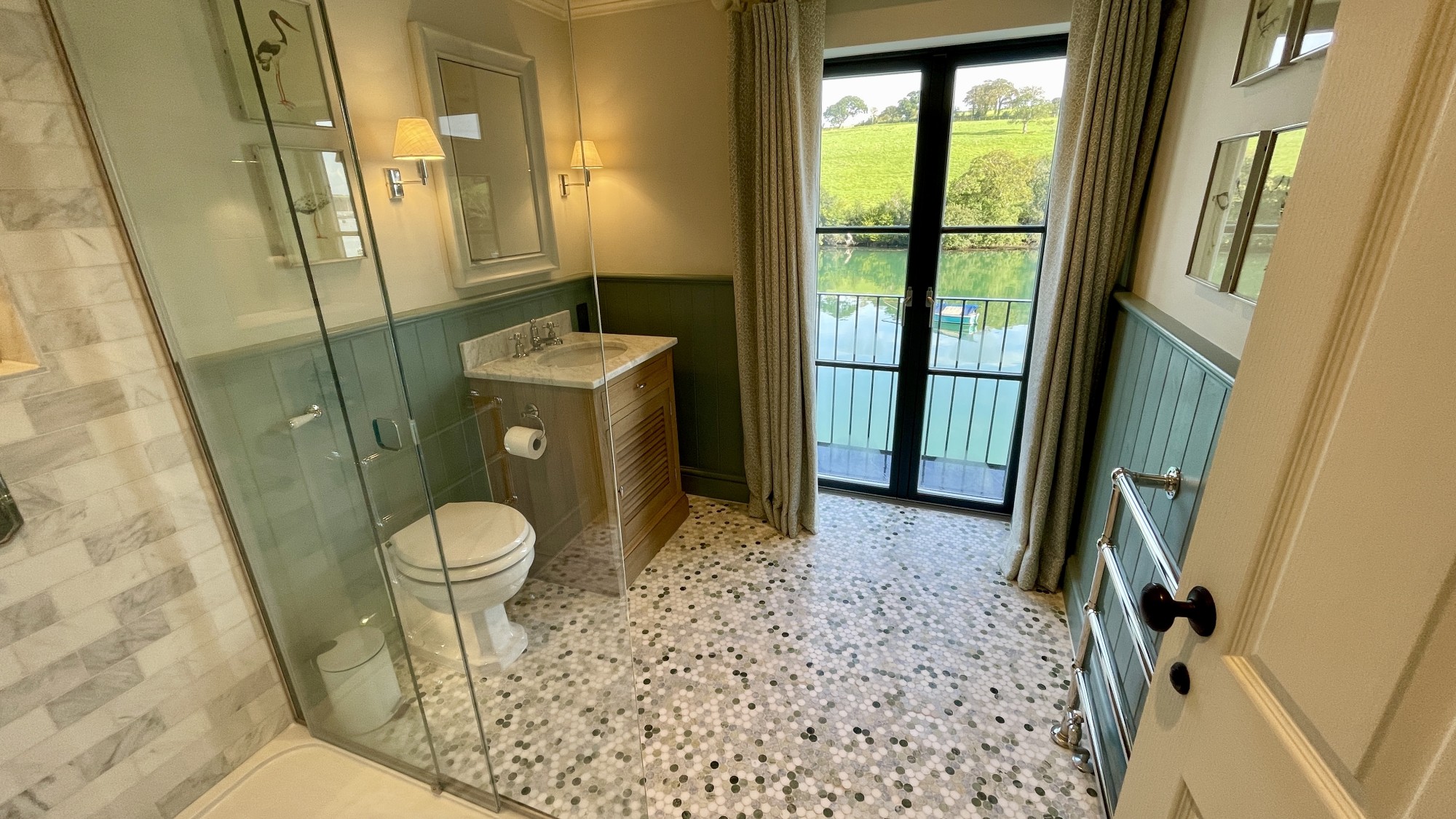 bathroom, with a glass-sided corner shower, toilet, basin and a floor-length window showing a garden beyond. Floor tiled in grey & silver random mosaic.