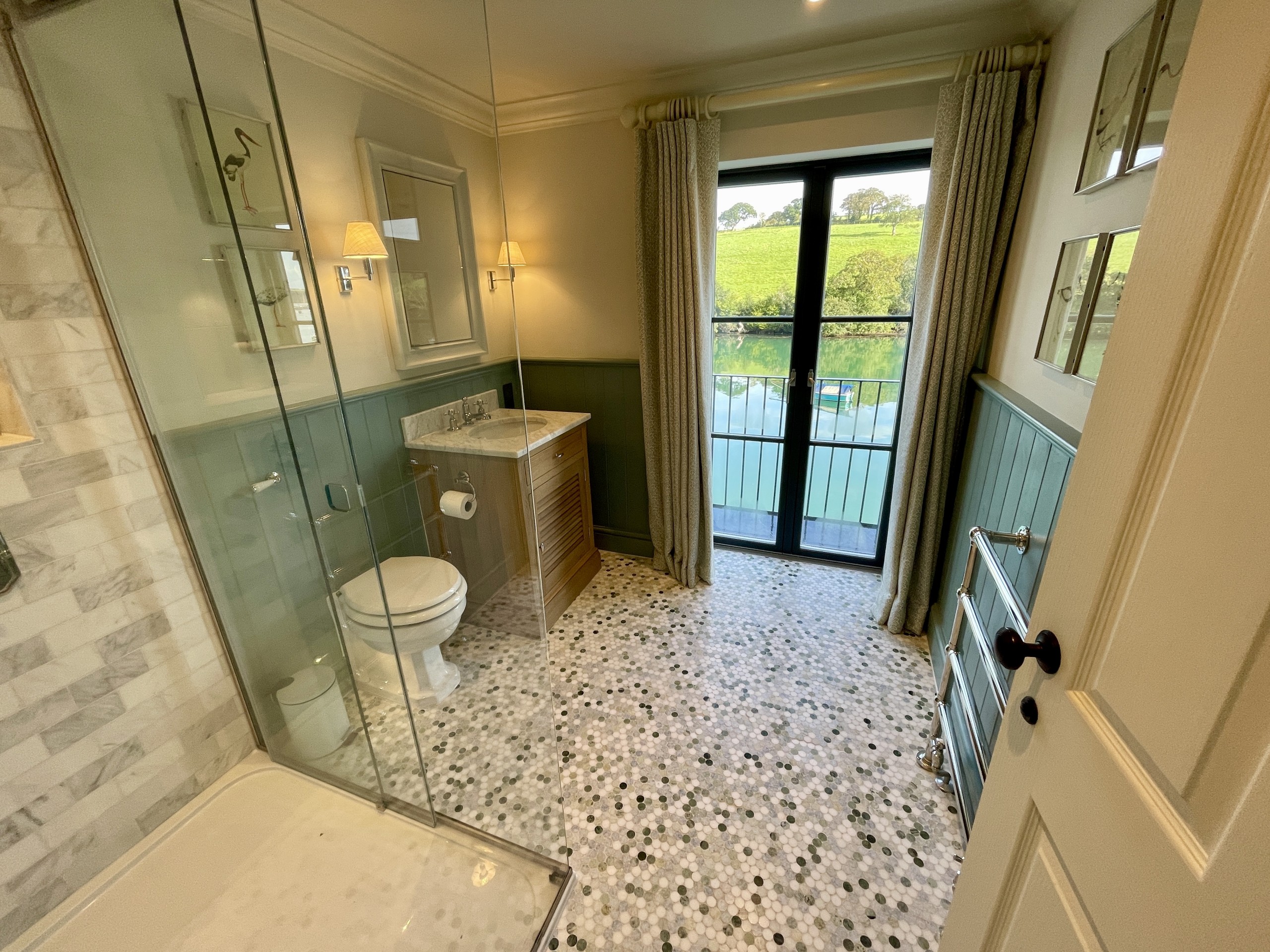 bathroom, with a glass-sided corner shower, toilet, basin and a floor-length window showing a garden beyond. Floor tiled in grey & silver random mosaic.